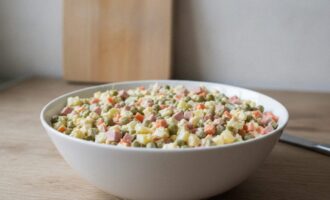 Салат оливье классический с колбасой в белой миске на кухонном столе